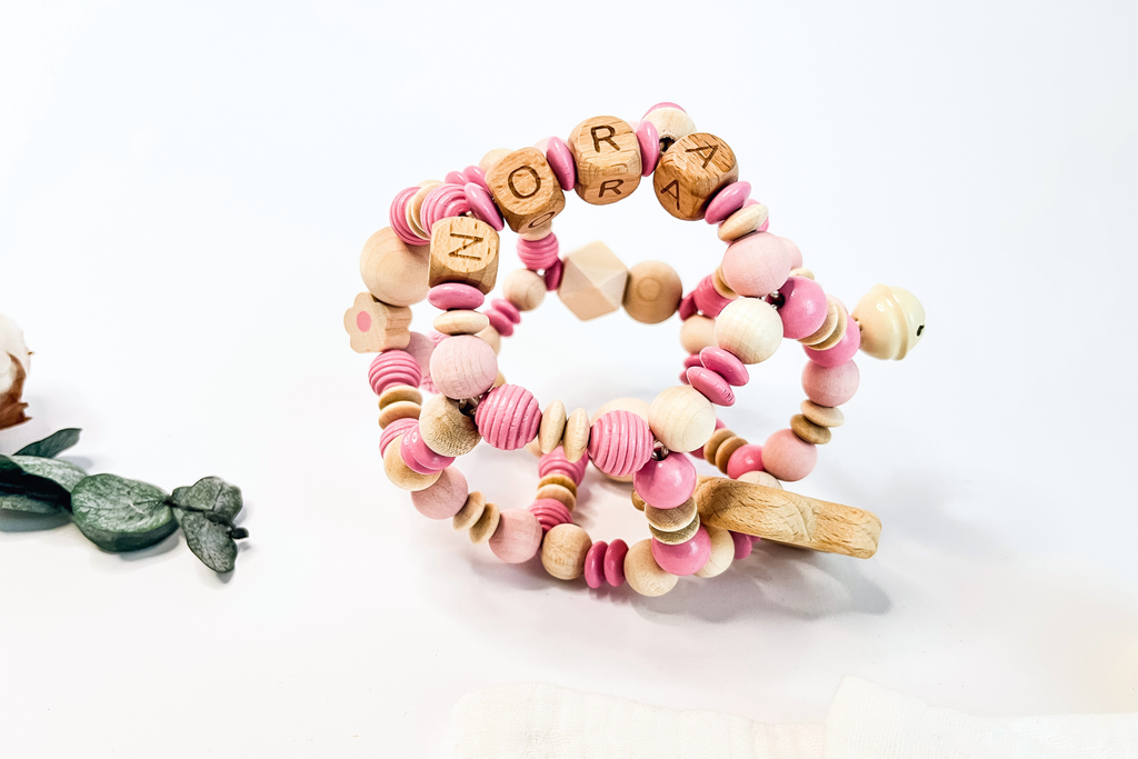 Personalisierter Motorikball aus Holz mit dem Namen NORA, in der Farbe Pink „Pink Peony“ mit dekorativer Holzfigur in Blumenform.